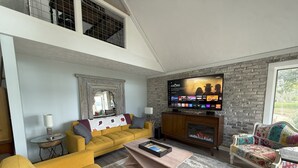 A living room with a yellow sofa, a fireplace, a TV, and a large window. - Three-bedroom cottage on Little Glen Lake (Empire)