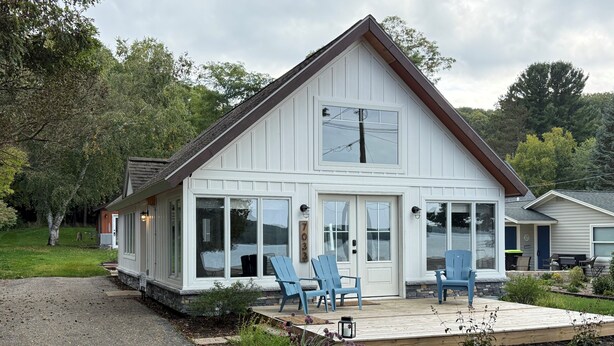 A white building with a gabled roof, large windows, and a deck with blue chairs. - Three-bedroom cottage on Little Glen Lake (Empire)