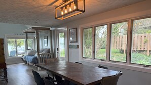 A spacious dining area with a long wooden table and modern chairs, large windows offering a view of the backyard, and a cozy seating area with a bench and cushions. - Three-bedroom cottage on Little Glen Lake (Empire)