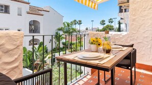 Un balcon avec une table en bois dressée pour le repas, une marquise jaune à rayures et une vue sur des palmiers et des immeubles.