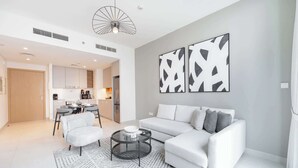 A modern living room with a white sofa, a round coffee table, and a patterned rug. There is a dining area with a table and chairs visible in the background.