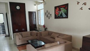 A living room with a beige sectional sofa, a glass coffee table, a clock on the wall, and a painting. - Hotel O Hari Villa (Jaipur)