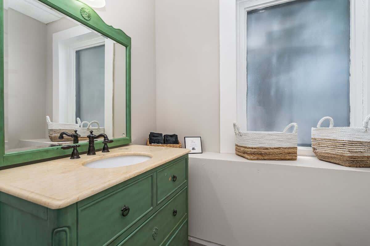 Une salle de bain avec un meuble-lavabo vert, un grand miroir, une fenêtre avec des paniers posés sur le rebord, et un lavabo blanc.