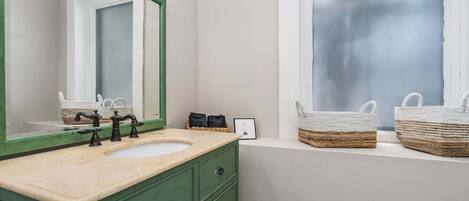 Une salle de bain avec un meuble-lavabo vert, un grand miroir, une fenêtre avec des paniers posés sur le rebord, et un lavabo blanc.