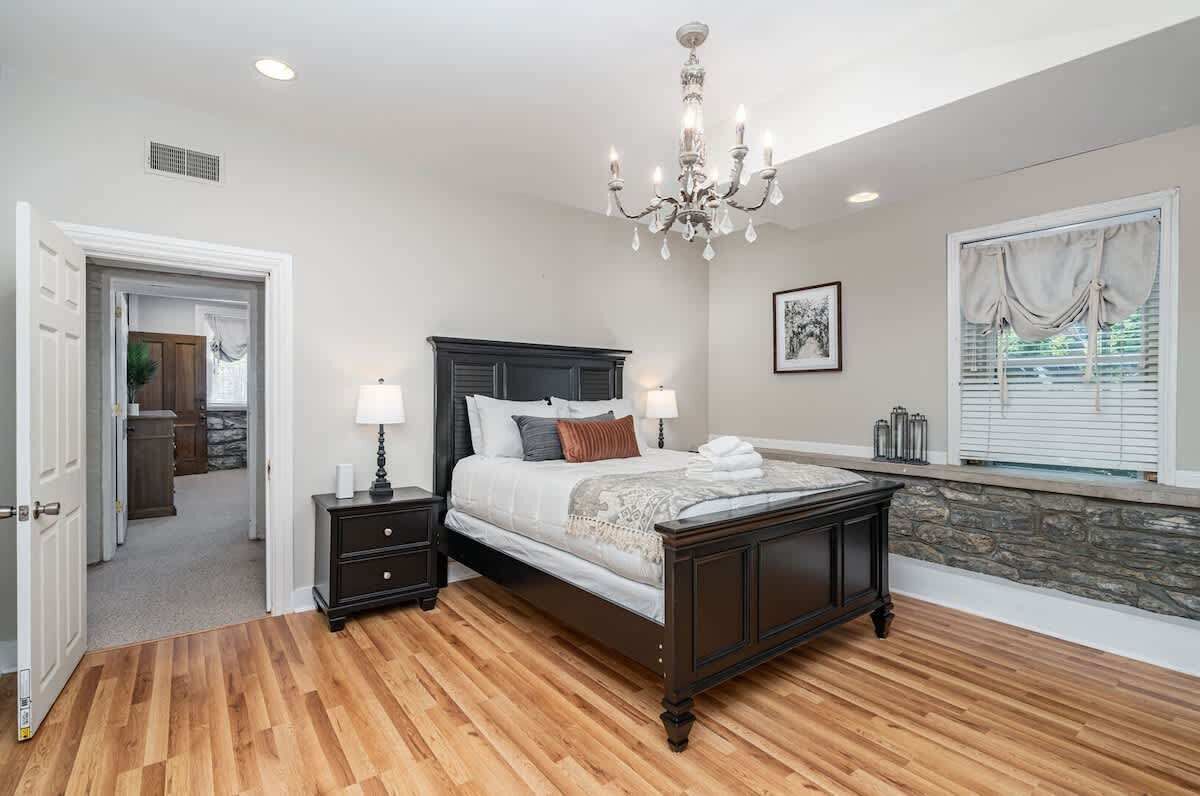 A bedroom with a large bed, a chandelier, a stone accent wall, a window with blinds, and a door leading to another room.