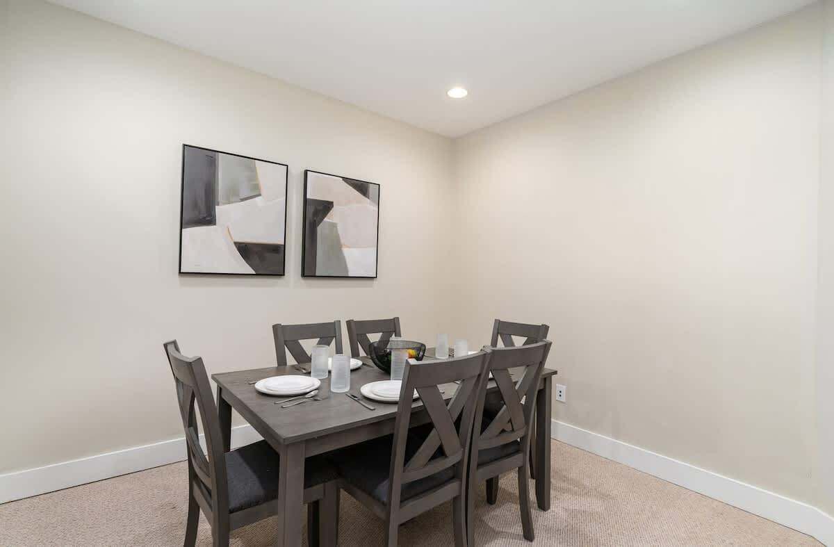 A dining area with a table set for four, four chairs, two abstract paintings on the wall, and a recessed ceiling light.
