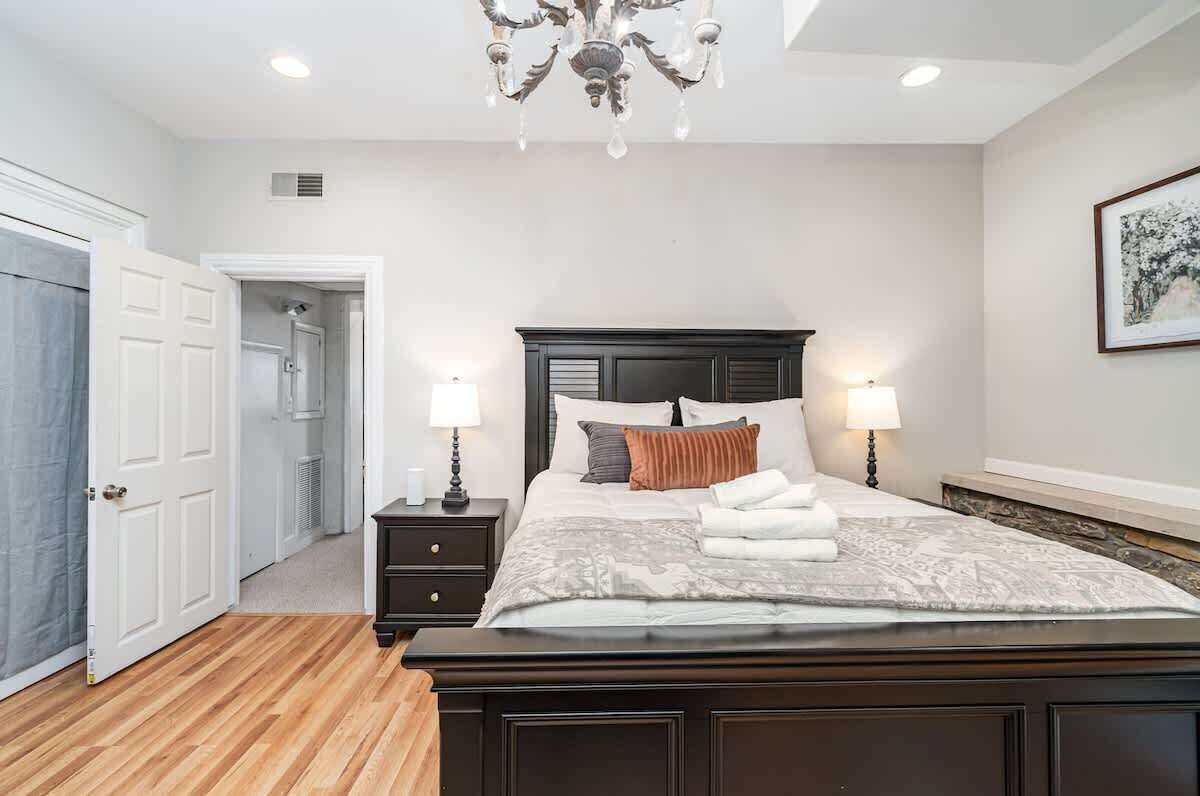 A bedroom with a dark wooden bed, two bedside tables with lamps, a chandelier, and a door leading to a hallway.
