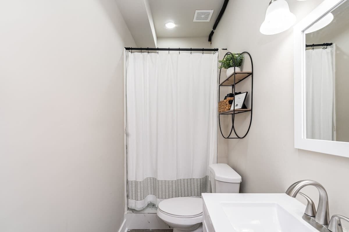 A bathroom with a white toilet, a white sink, a shower curtain, and a shelf with a plant and some items.