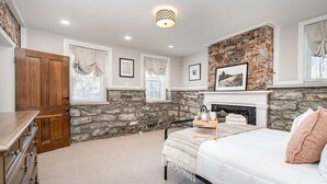A bedroom with a stone wall, a fireplace, a bed, a dresser, and a window with curtains.