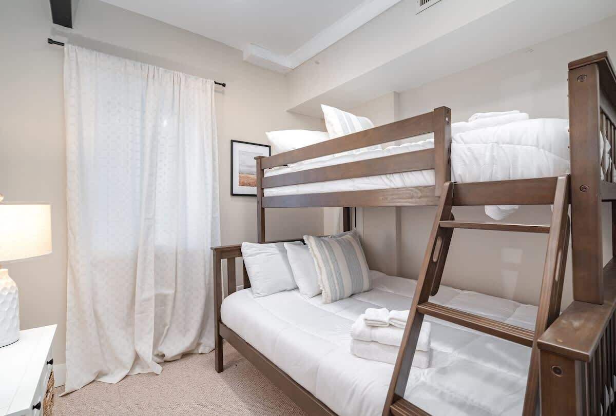 A bunk bed room with a wooden ladder, white bedding, a bedside table, and a curtain.