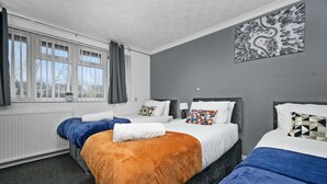 A hotel room with three beds, a large window with curtains, a radiator, and a wall-mounted artwork.