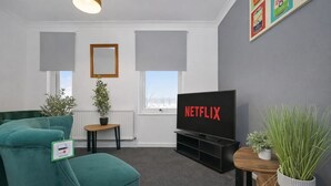 A modern living room with a teal sofa, a small wooden table, a potted plant, a black entertainment unit with a TV displaying 'NETFLIX,' and a framed picture on the wall.