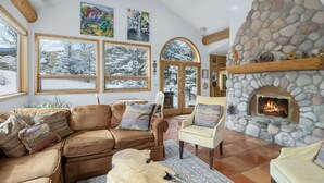 A cozy living room with a stone fireplace, large windows with snowy views, and a rustic wooden ceiling. - Aspen - Casa Connable (Aspen)