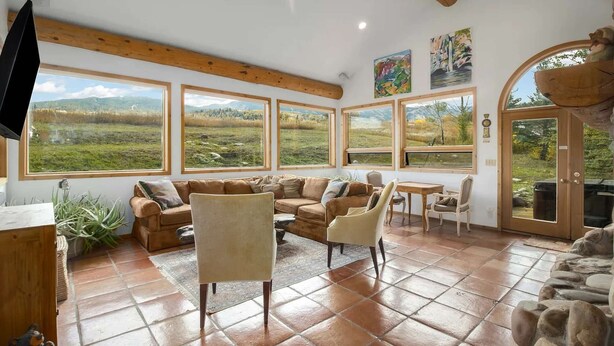A cozy living room with a large window overlooking a grassy field, a wooden ceiling, and a stone fireplace. - Aspen - Casa Connable (Aspen)