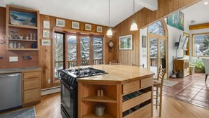 A modern kitchen with wooden cabinetry, a central island, and large windows offering a view of snow-covered trees. - Aspen - Casa Connable (Aspen)