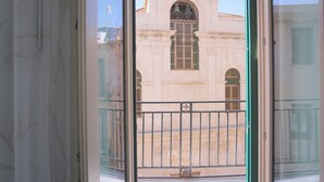 Une vue à travers une fenêtre ornée d’une rambarde en métal, donnant sur un bâtiment de style architectural classique, caractérisé par des fenêtres en arc et un fronton central.