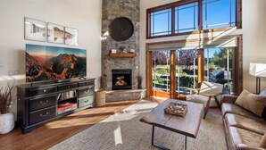A spacious living room with a stone fireplace, a large TV, and a view of the outdoors through large windows.