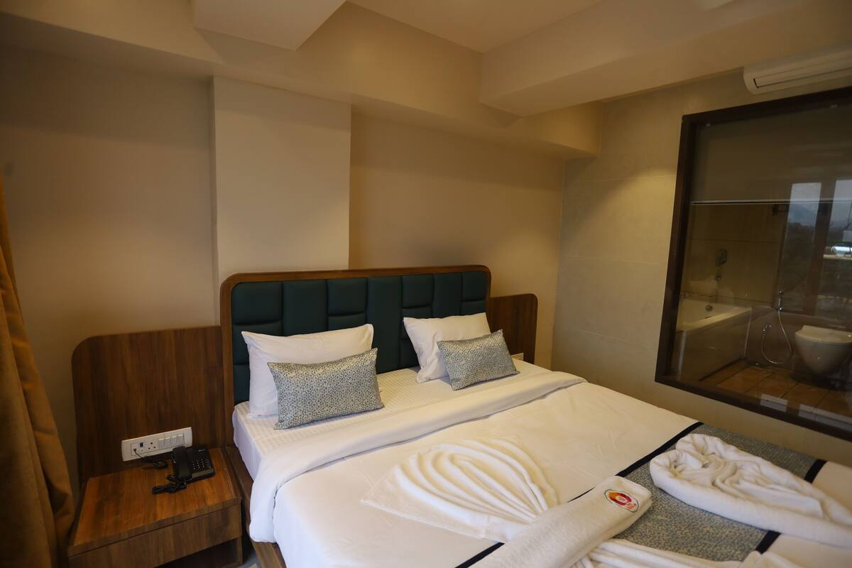 A hotel room with a neatly made bed, a bedside table with a phone, and a window with a view of a bathroom.