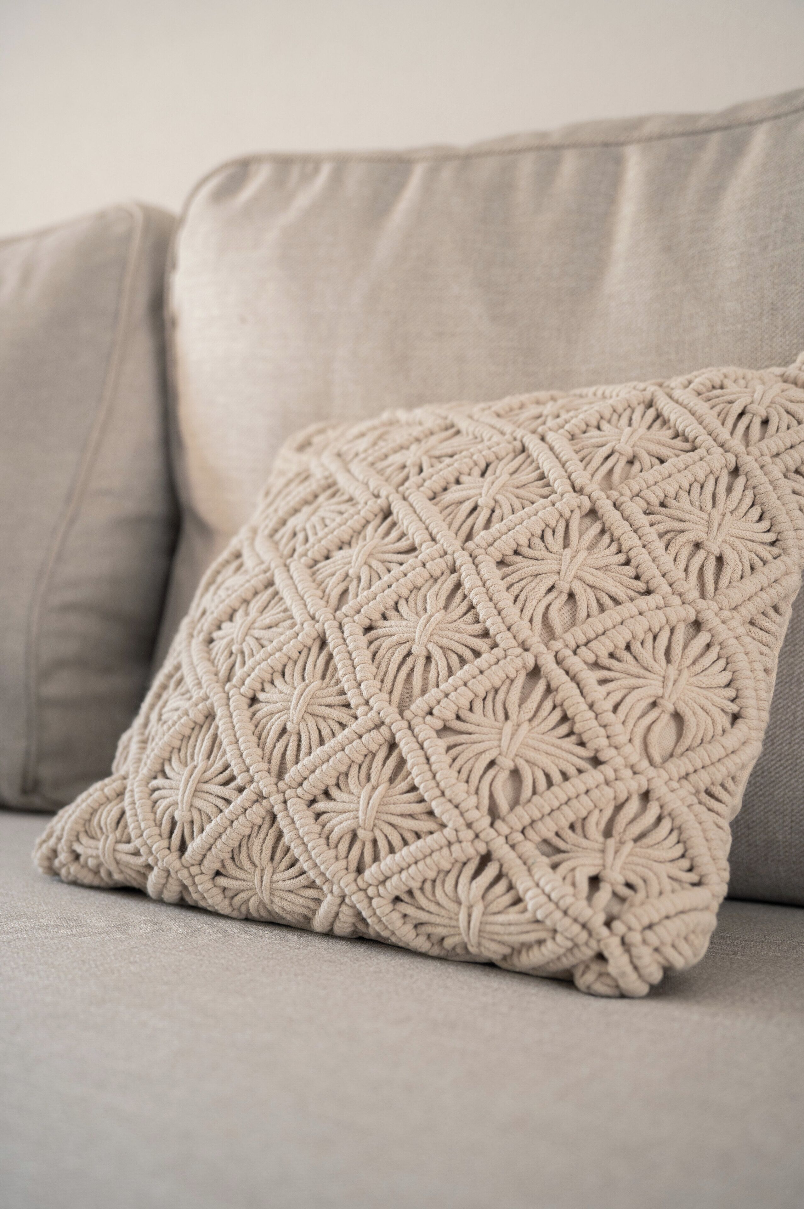 Un coussin en macramé de couleur beige, posé sur un canapé gris clair.