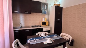 Een kleine keuken met een gasfornuis, magnetron en kastjes. Er is een eettafel met stoelen en een tafelkleed.