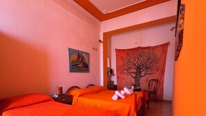 Een kamer met twee bedden belegd met oranje dekens, een eettafel met stoelen, een wandkleed met een ontwerp van een boom, en een schilderij aan de muur.