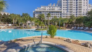 Una piscina con sillones de descanso y palmeras, rodeada por un edificio hotelero de varios pisos.