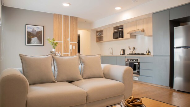 Ein modernes Wohnzimmer mit einem beigefarbenen Sofa, einem Holztisch und einer Küchenzeile mit hellblauen Schränken und Edelstahlgeräten.