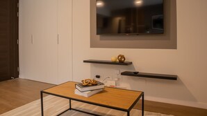 Ein modernes Wohnzimmer mit einem an der Wand montierten Flachbildfernseher, einem Holztisch mit Büchern und einem Brezel sowie einem kleinen Regal mit Dekoration.