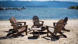 A beach with three wooden Adirondack chairs and a fire pit, overlooking a lake with boats and mountains in the background. - Fresh Design, King & Queen - Seasons at Sandpoint (Sandpoint)