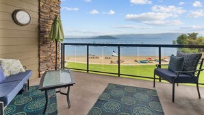 A balcony with a view of a beach, a clock, and outdoor furniture. - Fresh Design, King & Queen - Seasons at Sandpoint (Sandpoint)
