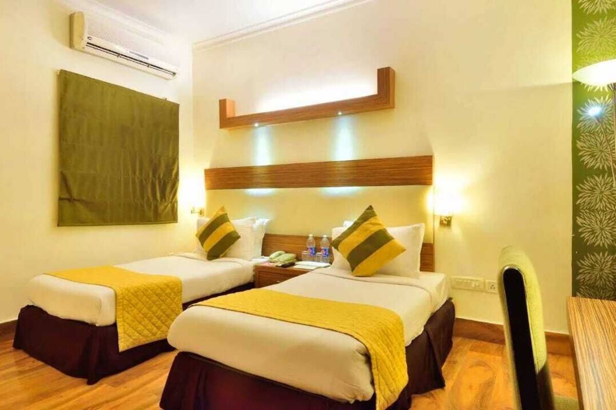 A hotel room with two beds, a wooden floor, a green wall, and a ceiling-mounted air conditioner.