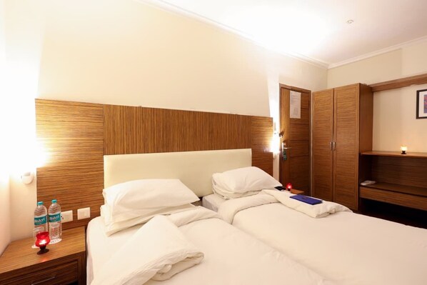 A hotel room with a wooden headboard, a bed with white linens, a nightstand with water bottles, a bedside lamp, and a wardrobe. - Sk Premium Park GGN (Gurugram)