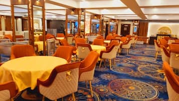 Uma sala de jantar de hotel com mesas redondas, cadeiras laranja e um tapete estampado.
