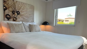 A neatly made bed with a white quilt, a large abstract wall art piece, and a window with a view of buildings outside. - Beautiful Country Home for 6 (Sint Maartensvlotbrug)