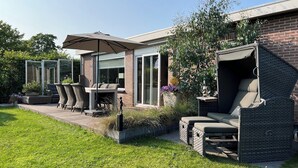 A modern outdoor patio with wicker furniture, a glass enclosure, and a brick house in the background. - Beautiful Country Home for 6 (Sint Maartensvlotbrug)