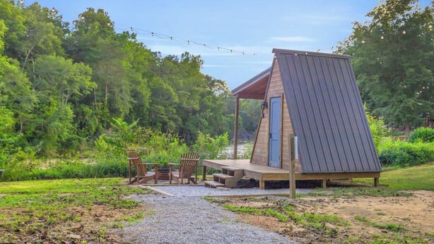 A small, triangular wooden structure with a porch and seating area, surrounded by greenery and a gravel path. - The Camellia - A Frame Camping Pod w/ River View (Easley)