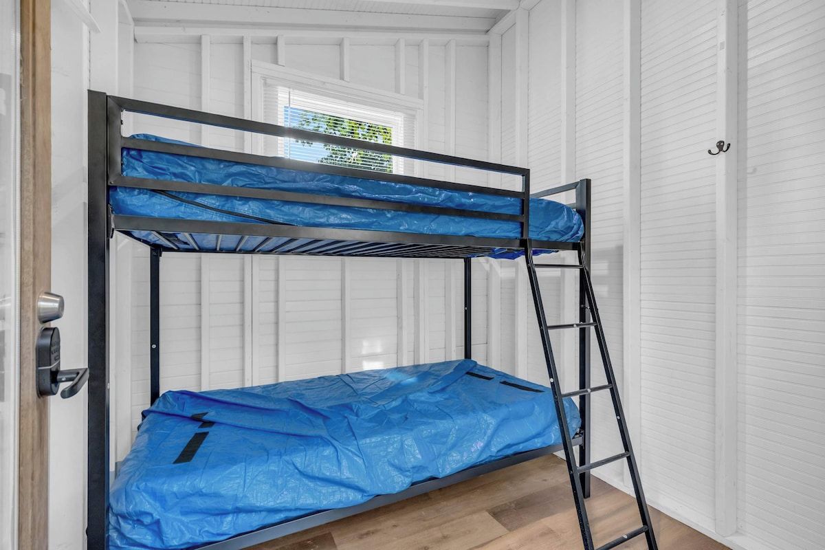 Una cama litera con un cubrecolchón azul, una escalera metálica y una pared blanca con paneles.