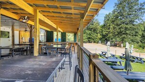 An outdoor covered patio with wooden tables and chairs, a barbecue area, and picnic tables with umbrellas. - The Daffodil - Cozy Camping Pod w/ River View (Easley)