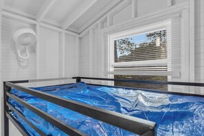 A room with a bed covered in blue sheets, a window with blinds, and a wall-mounted fan. - The Daffodil - Cozy Camping Pod w/ River View (Easley)