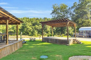 A covered outdoor area with a wooden deck, picnic tables, and a building in the background. - The Jessamine - Riverview Camping Pod, Close to DT (Easley)