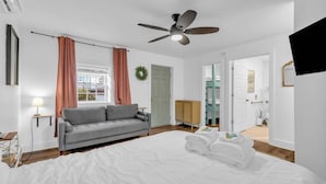 Una habitación de hotel con una cama, un sofá gris, un ventilador de techo, un televisor y una puerta que da a otra habitación.