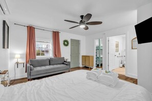 A hotel room with a bed, a grey sofa, a ceiling fan, a TV, and a door leading to another room.