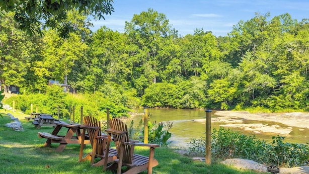 Un espacio de picnic con sillas y mesas de madera, un río y un paisaje boscoso de fondo.