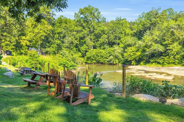 A picnic area with wooden chairs and tables, a river, and a forested backdrop.