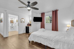 A bedroom with a bed, a ceiling fan, a TV, a window with curtains, and a bathroom with a sink and mirror.