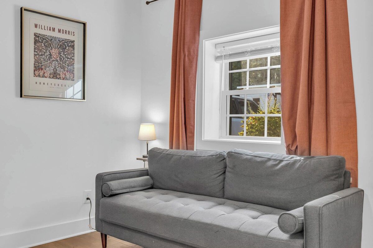 A grey sofa in a room with a window, a lamp, and a framed picture on the wall.