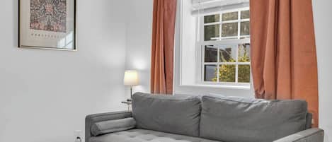 Un sofá gris en una habitación con una ventana, una lámpara y un cuadro en la pared.
