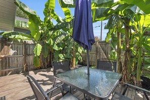 A glass-top patio table with metal chairs, surrounded by banana plants and a wooden fence.