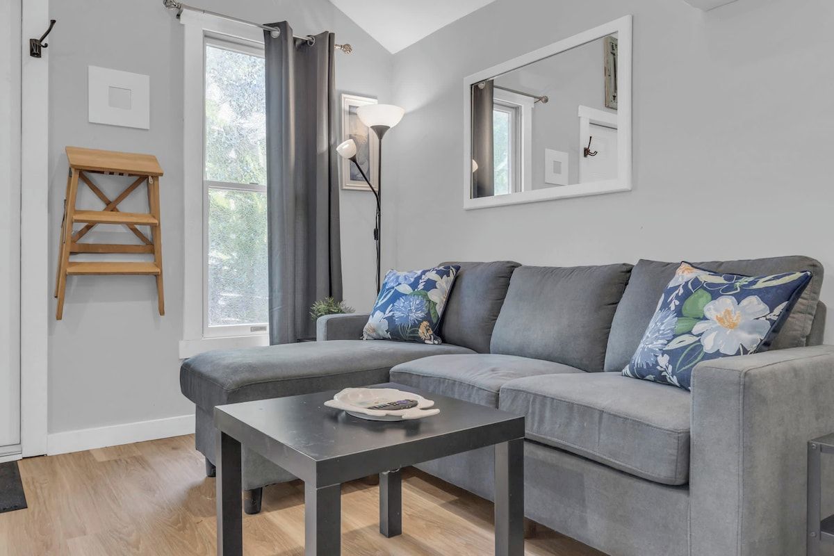 Una sala de estar moderna con un sofá gris, una mesita, una escalera de madera y un ventanal con cortinas.