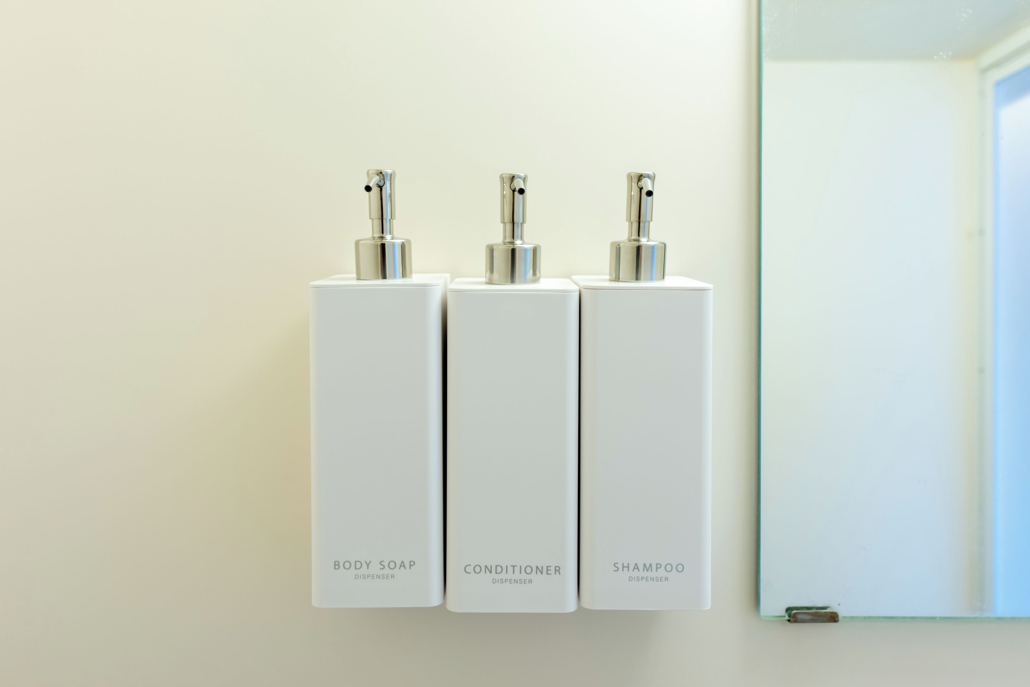 Tres dispensadores blancos etiquetados como 'BODY SOAP', 'CONDITIONER' y 'SHAMPOO' montados en una pared.